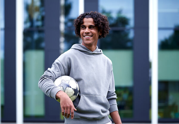 A young man in a grey hoodie, smiling and holding a football under his arm.