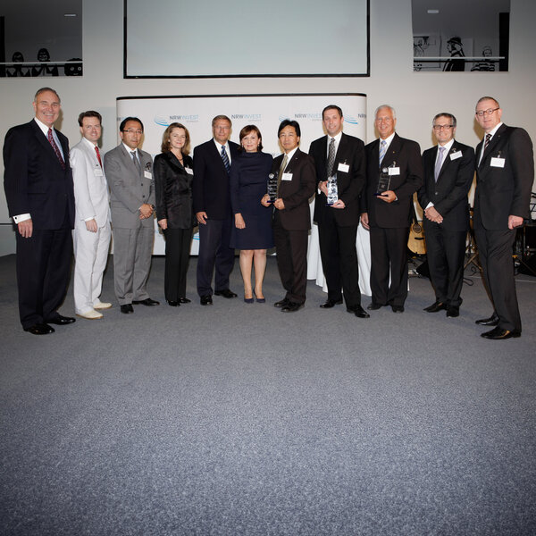 Twelve people in business attire stand side by side, three holding glass trophies, with a screen bearing the words “NRW Invest Germany” in the background.