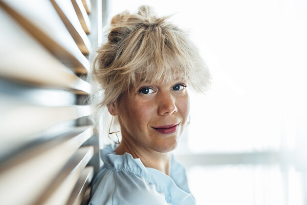 A portrait photograph of a woman leaning against an office wall wearing a light blue blouse.