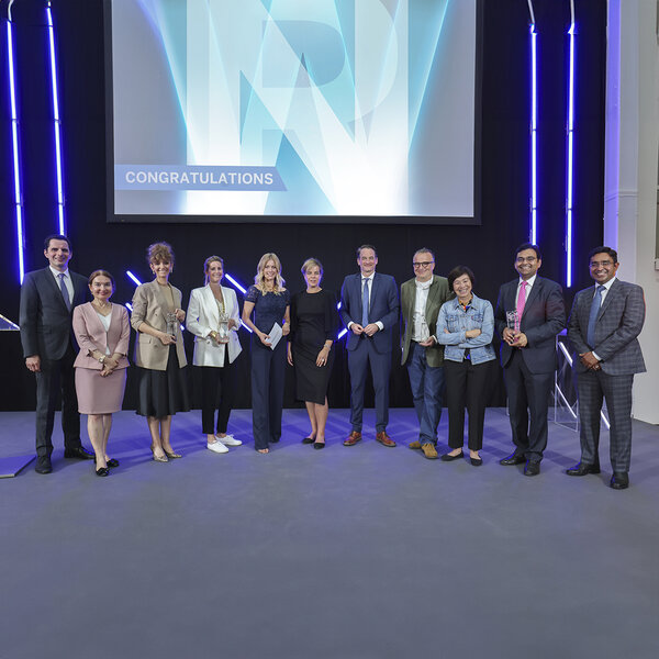 Eleven people in business attire stand in a group, some holding glass trophies, with the word “Congratulations” displayed on a large monitor behind them.