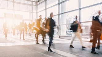 Blurred photograph of several people in motion as they enter or leave a light-filled hall with a glass front.