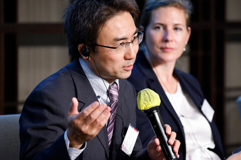 A man in business attire speaks into a microphone. Next to him sits a woman, also dressed in business attire.