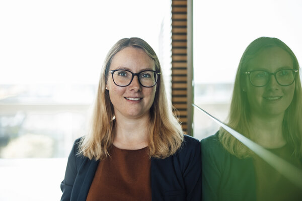 A portrait photograph of a woman in front of an office window. She is wearing glasses and a dark blue blazer.