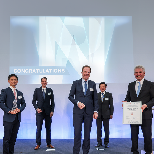 Five men in suits are standing on a stage, one holding a certificate, another holding an award. On the screen behind them is the word “Congratulations”.