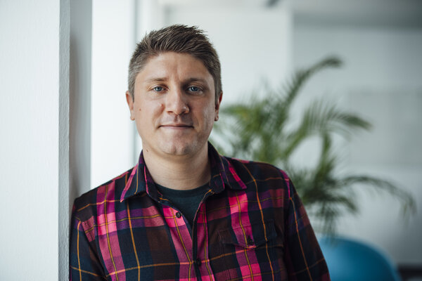 A portrait photo of a man with short, styled brown hair. He is wearing a checked shirt and leaning sideways against an office wall.