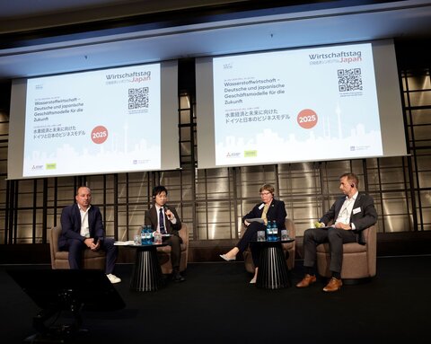 Four people in business attire are seated on a stage in armchairs, behind them projections with the words “Japan Business Day 2025”.