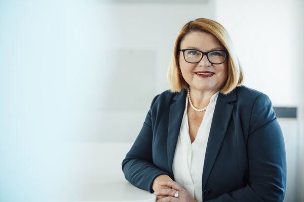 A portrait photograph of a smartly dressed woman. She is wearing glasses and a pearl necklace around her neck.