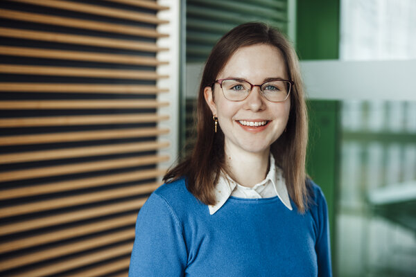 Portrait photograph of a young woman. She is wearing glasses and a blue sweatshirt over a light-coloured top.