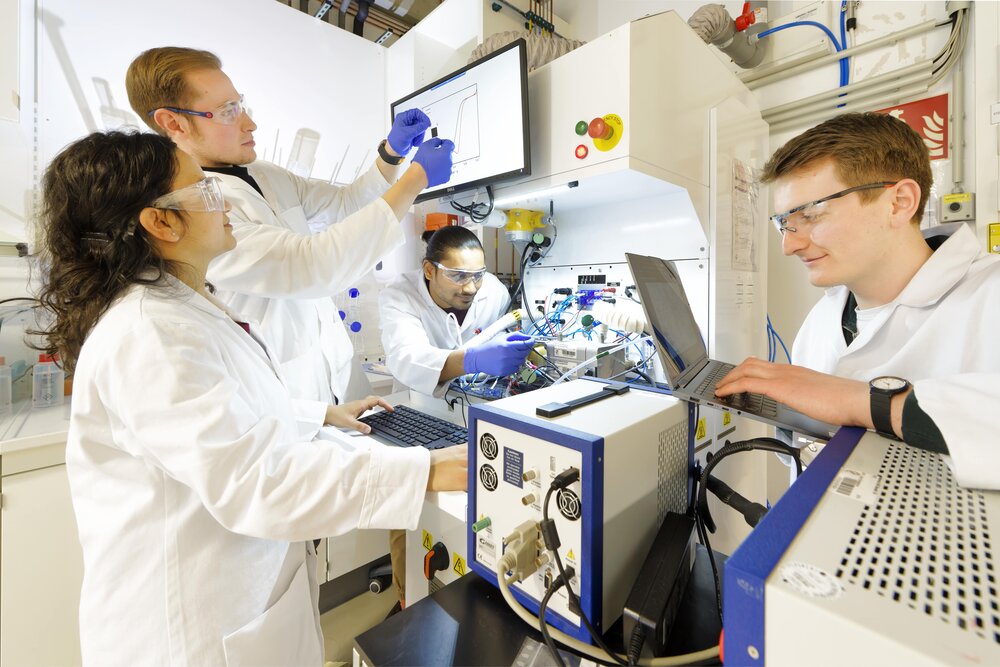 Vier Personen mit Schutzbrillen und in Laborkitteln arbeiten gemeinsam an einem Versuchsaufbau und schauen auf die Displays von einem Laptop und Monitor.