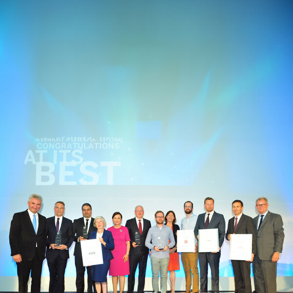 Twelve people in business attire stand in a group, some holding certificates and glass trophies, with the projection Congratulations at its best on the wall behind them.