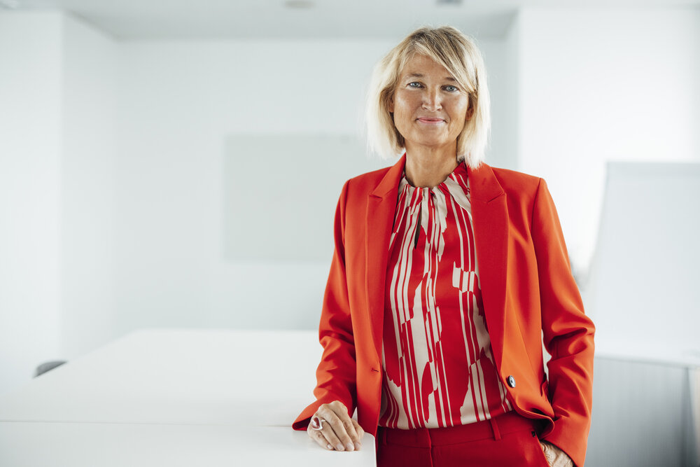 A portrait photograph of a smartly dressed woman wearing various orange garments. She is standing next to an office desk with her arm resting on it.