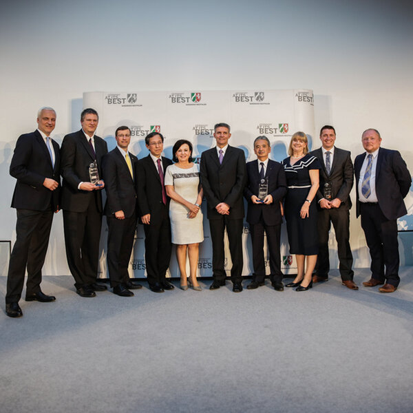 Ten people in business attire stand next to each other, several holding glass trophies, with a screen in the background displaying the words “Germany at its best”.