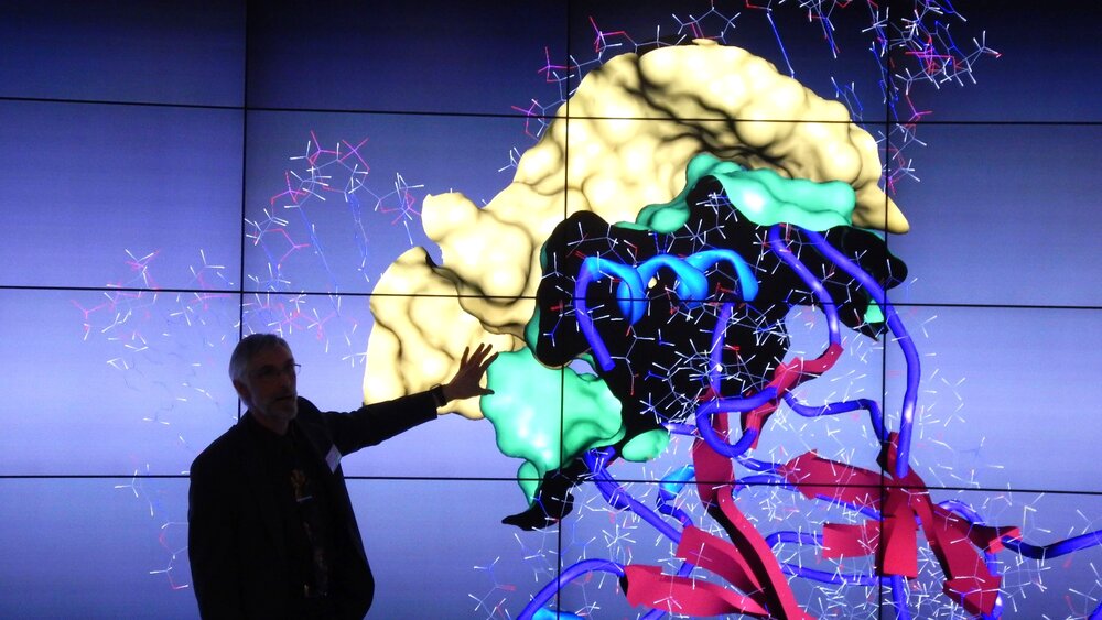 Ein Mann präsentiert auf einer großen Leinwand ein 3D-Modell von Molekülstrukturen mit bunten grafischen Elementen.