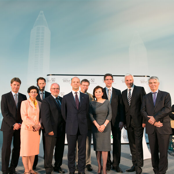 Ten people in business attire stand side by side, three holding glass trophies, with a backdrop featuring the words “Germany at its best” and stylised towers.