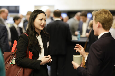 A woman and a man in business attire are conversing; she is carrying a red bag, he is holding a cup, blurred background with other people.