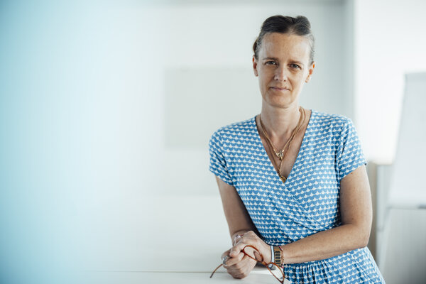 A portrait photo of a woman in a blue and white summer dress. She is standing at an office desk and holding her glasses in her hand.