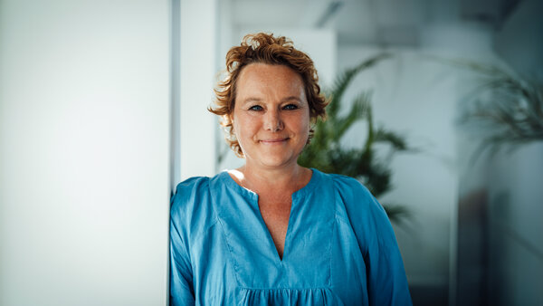 A portrait photograph of a woman wearing a blue blouse standing sideways against an office wall.