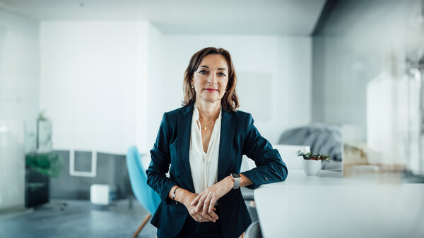 A portrait photo of a smartly dressed woman wearing various pieces of jewellery. She is standing sideways at an office desk with her arm resting on it.