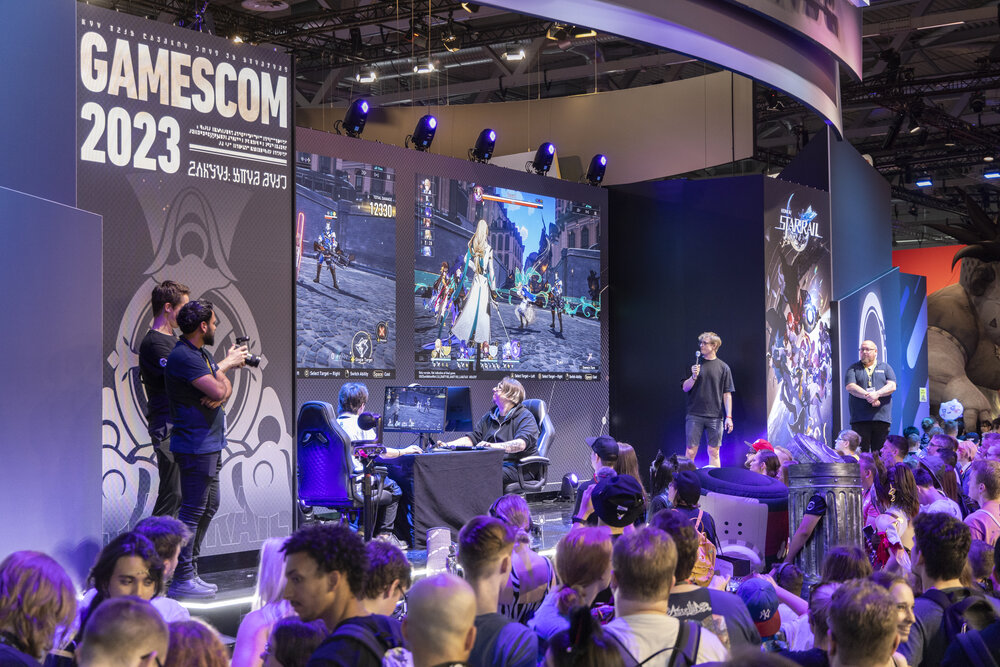 Eine Bühne der Gamescom 2023 auf der zwei Personen ein Computerspie spielen, was auf einer Leinwand vor Zuschauern übertragen wird.