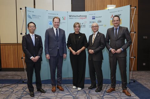 Five people in business attire stand in front of a wall with logos and lettering.