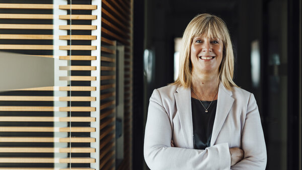 A portrait photograph of a smartly dressed woman. She has her arms crossed and is wearing a light grey blazer and a necklace.
