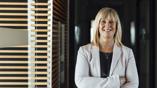 A portrait photograph of a smartly dressed woman. She has her arms crossed and is wearing a light grey blazer and a necklace.