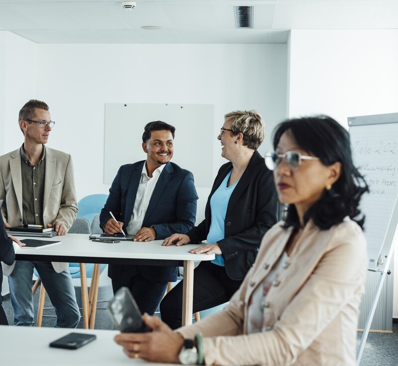 Several people in business attire are sitting and standing in a bright meeting room, with a flipchart containing notes in the background.