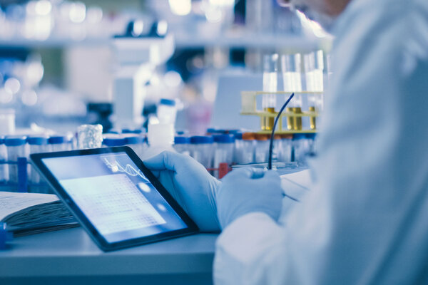 Eine Person im Labor arbeitet mit einem Tablet, umgeben von Reagenzgläsern und Notizen.