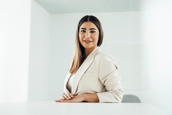 A portrait photo of a young woman wearing a beige blazer. She is sitting sideways at an office desk with her hands resting on it.