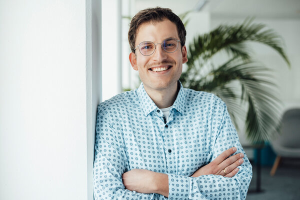 A portrait photograph of a man leaning sideways against an office wall. He is wearing glasses and a light-coloured patterned shirt. 