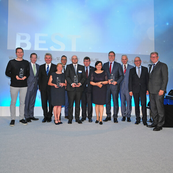 Twelve people in business attire stand side by side, several holding glass trophies, with a screen in the background displaying the words “Germany at its best”.
