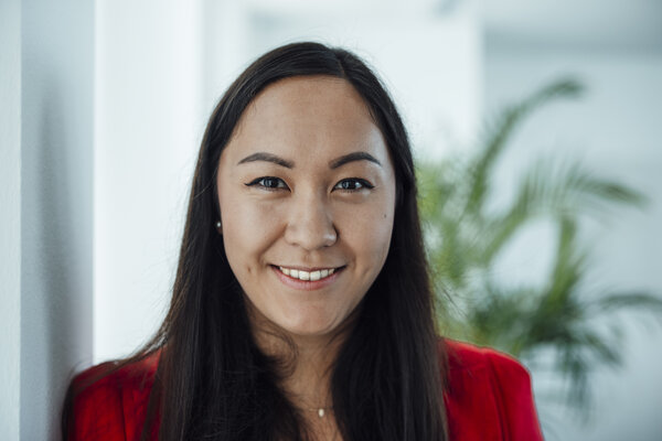 A portrait photograph of a woman standing against an office wall wearing a reddish top.