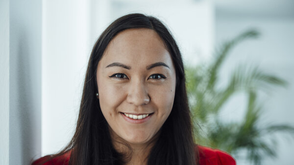 A portrait photograph of a woman standing against an office wall wearing a reddish top.