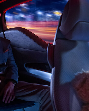 A man is sitting in the back seat of a car, holding a tablet; next to him, a teddy bear lies on the front seat; outside, lights are passing by in the night.
