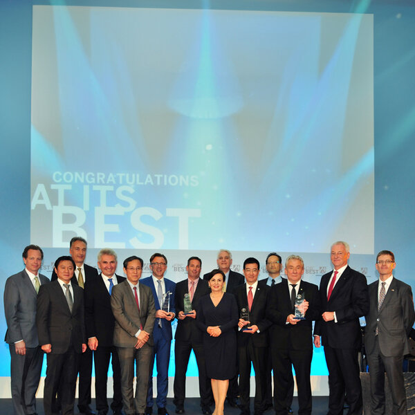 Fourteen people in business attire stand in a group, several holding glass trophies, with the projection Congratulations at its best on the wall behind them.